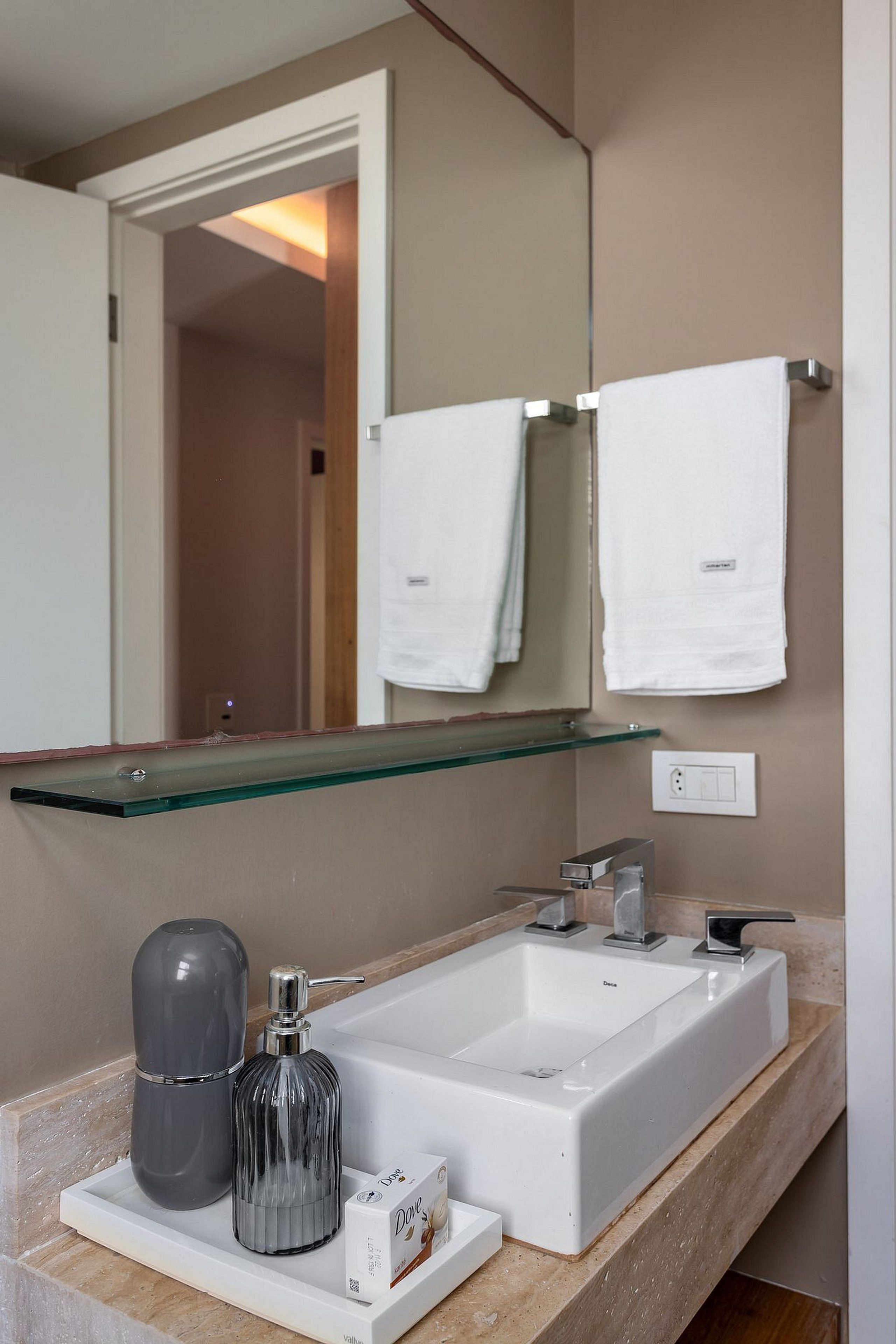 A modern bathroom with a large mirror, two white towels, a glass shelf, a white sink, and a soap dispenser.