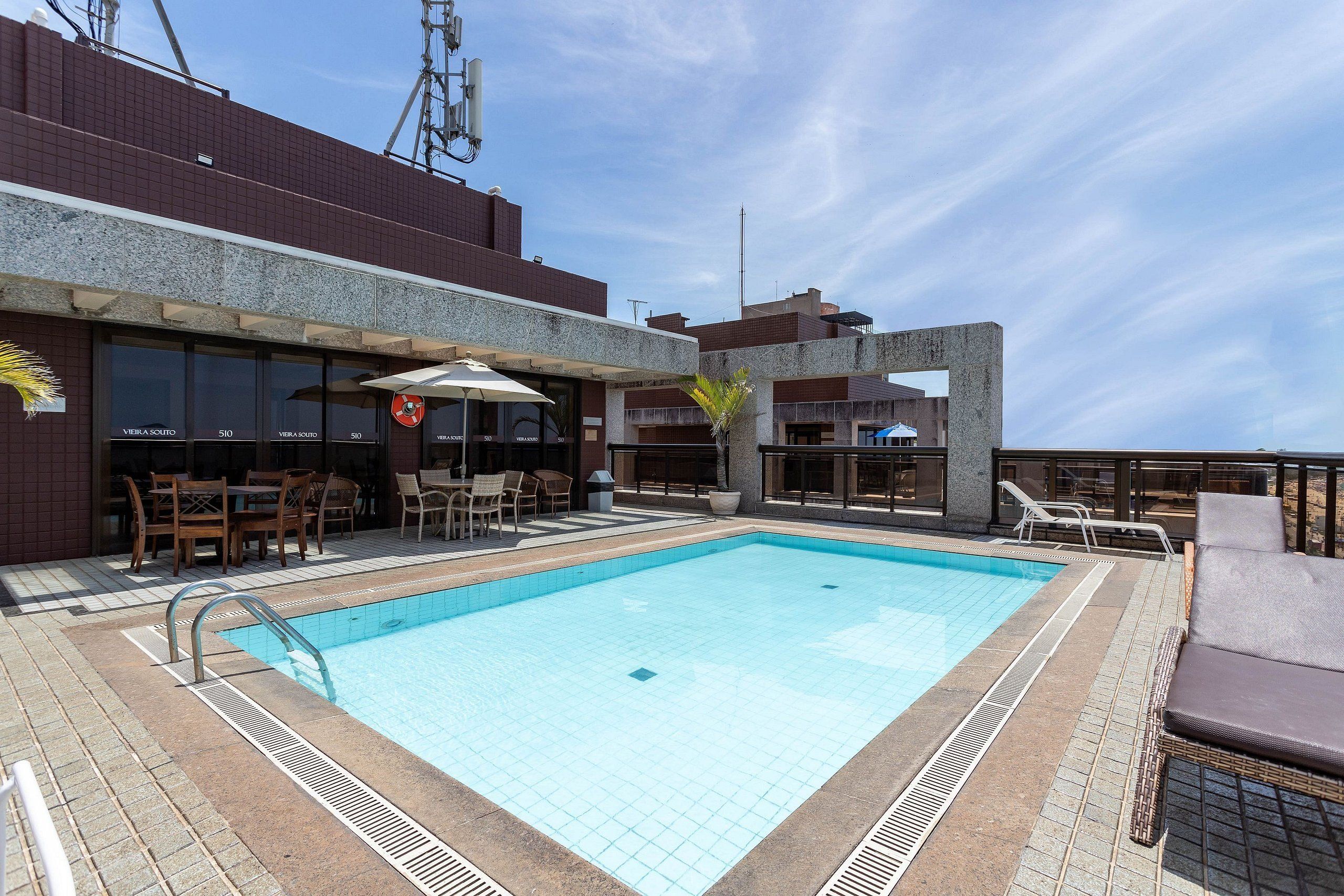 A rooftop pool with a clear blue water surface, surrounded by lounge chairs and a bar area with wicker furniture.