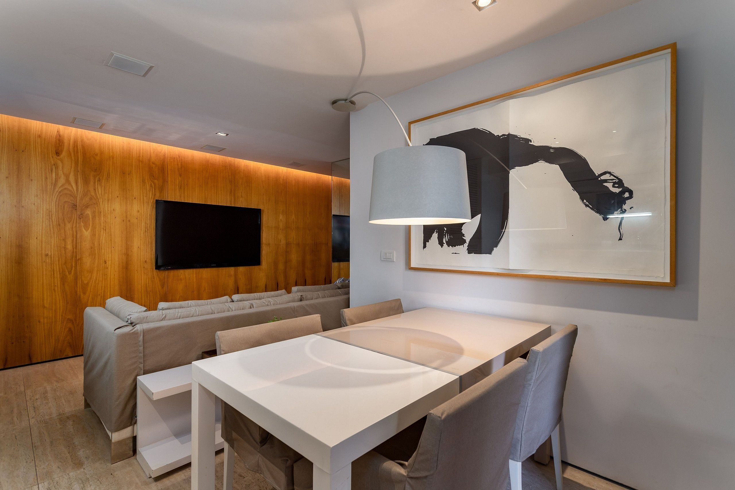 A modern living room with a wooden wall, a flat-screen TV, a grey sofa, a dining table, and a large abstract painting.