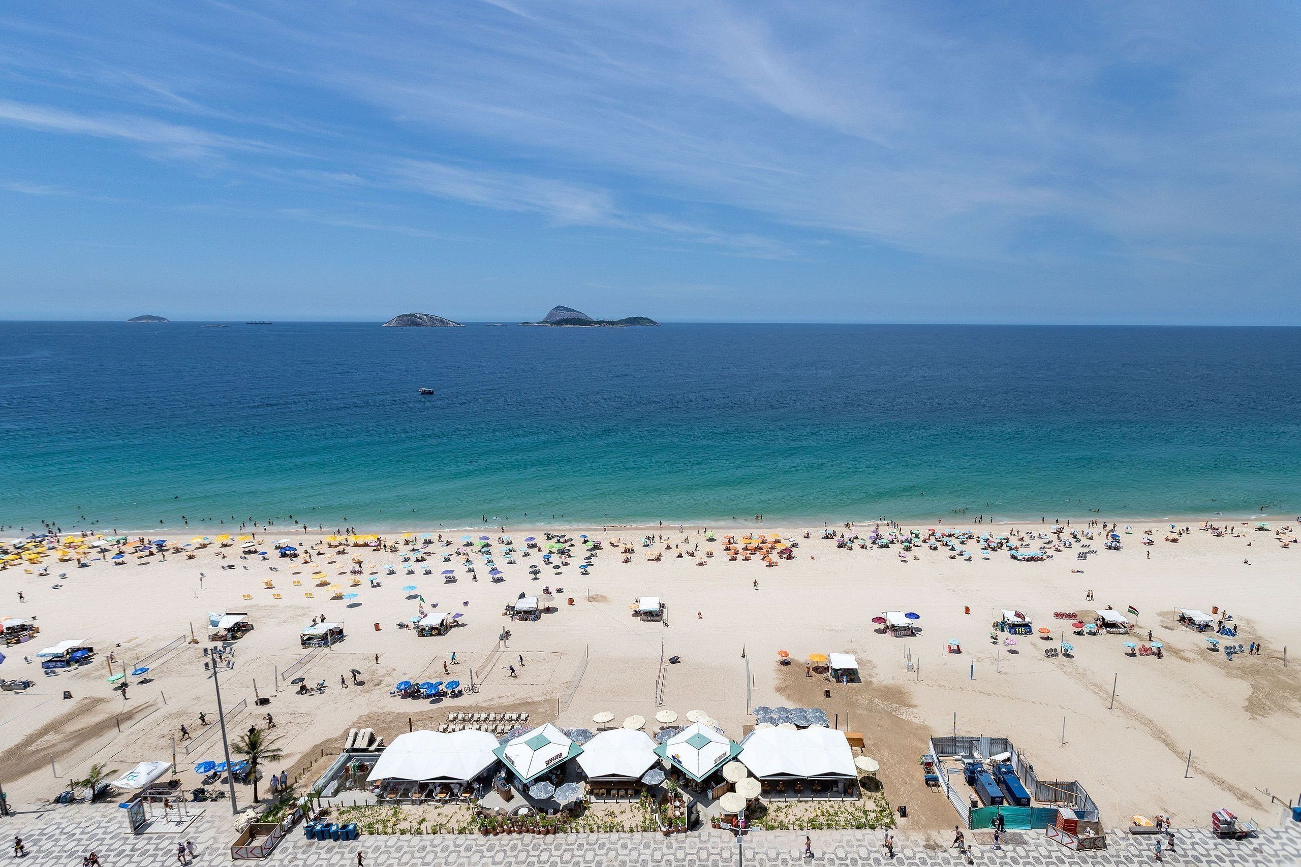 A beach with numerous umbrellas and people, a clear sky, and distant islands.