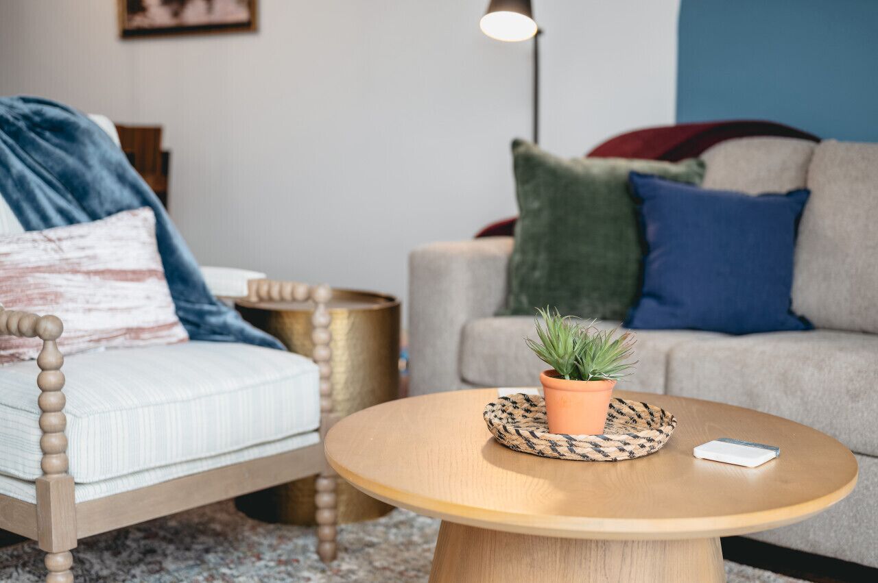 Ein gemütliches Wohnzimmer mit einem runden Holztisch, einer kleinen Topfpflanze und einem Smartphone darauf.