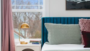Un dormitorio con cabecera azul, lámpara de noche, ventana con cortinas y algunos libros en la mesita de noche.