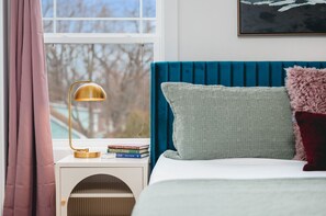 Un dormitorio con cabecera azul, lámpara de noche, ventana con cortinas y algunos libros en la mesita de noche.