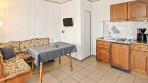 Um espaço de estar compacto com sofá, uma mesa pequena com toalha, televisão montada na parede, área de cozinha com armários de madeira e cafeteira, e piso revestido de azulejos.
