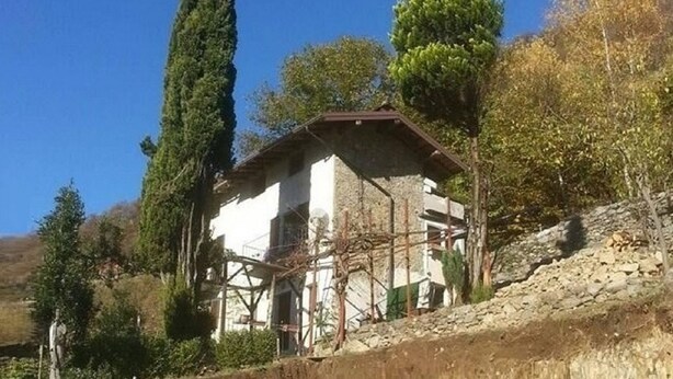 Um edifício rústico de pedra com telhado inclinado, rodeado por árvores altas e um céu azul límpido.