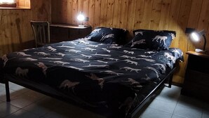 Quarto com paredes de madeira, cama com roupa de cama estampada, uma cadeira e duas luminárias de cabeceira.