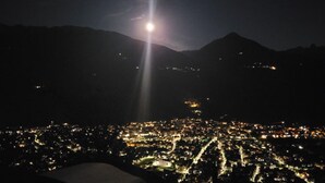 Paisagem urbana noturna com uma montanha imponente ao fundo e uma lua brilhante no céu.