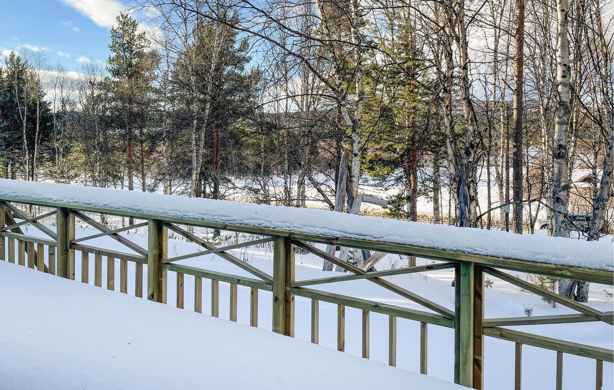 A snow-covered wooden deck with a metal railing, surrounded by trees and a clear sky.