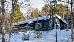 En hytte med sneklædt tag, omgivet af træer og en træterrasse.