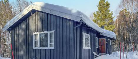 A small, dark wooden cabin with a snow-covered roof, surrounded by trees and a snowy landscape.