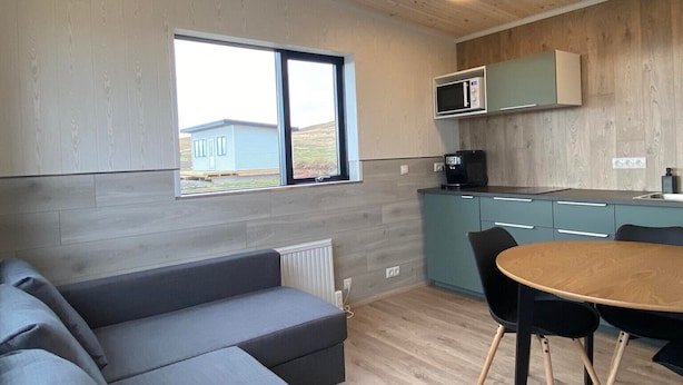 A modern interior with a grey sectional sofa, a wooden dining table with chairs, a kitchen area with a microwave, and a large window offering a view of the outdoors. - Brekkukot Cottage  (Blönduós)