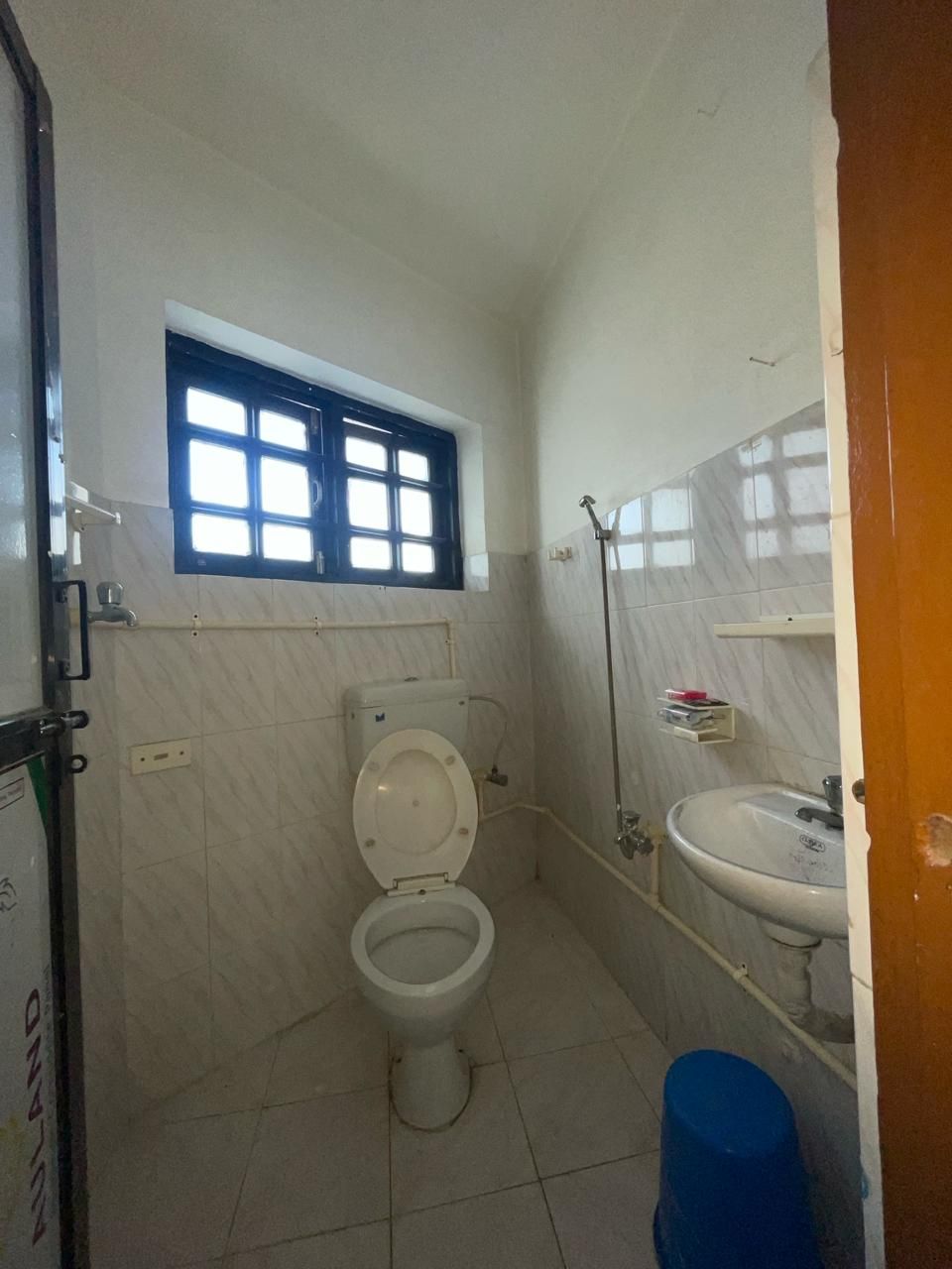 A small bathroom with a toilet, a window, a sink, and a blue trash bin.