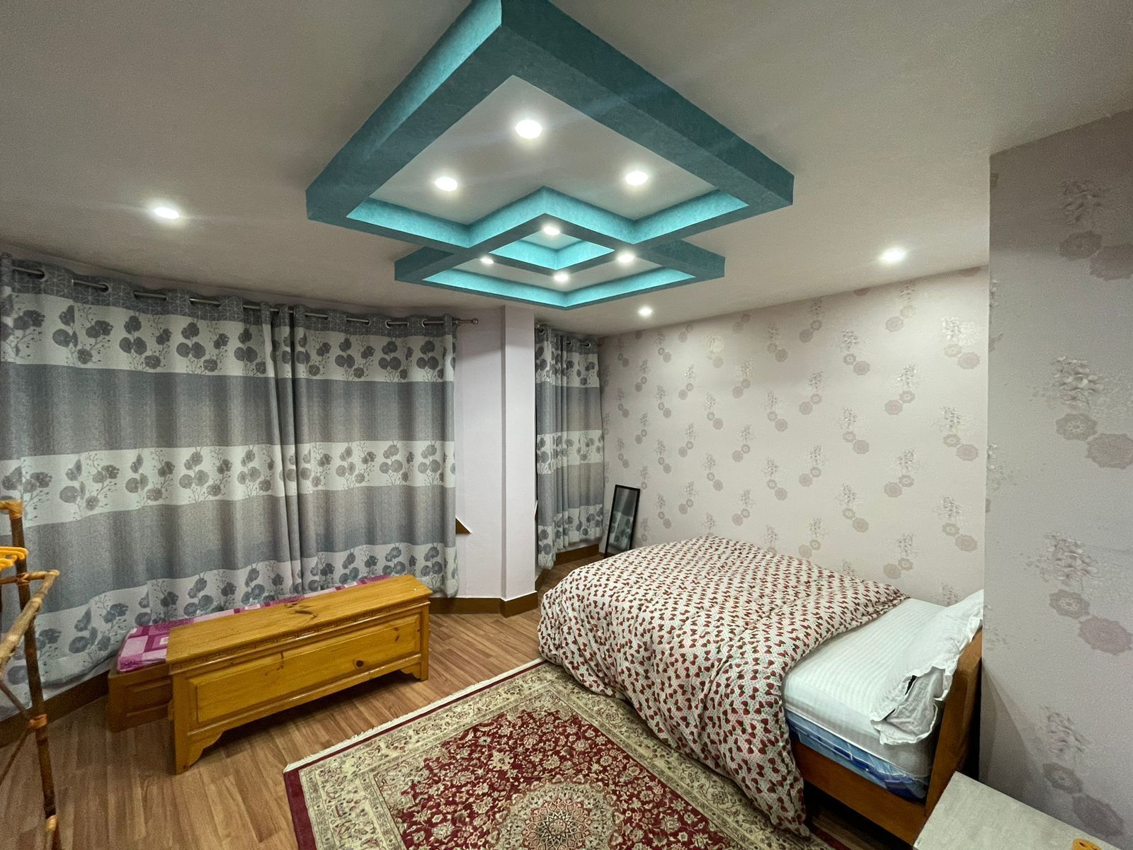 Une chambre à coucher comprenant un lit, une commode en bois, une table de chevet, un tapis à motifs et un plafond doté d’un éclairage encastré.