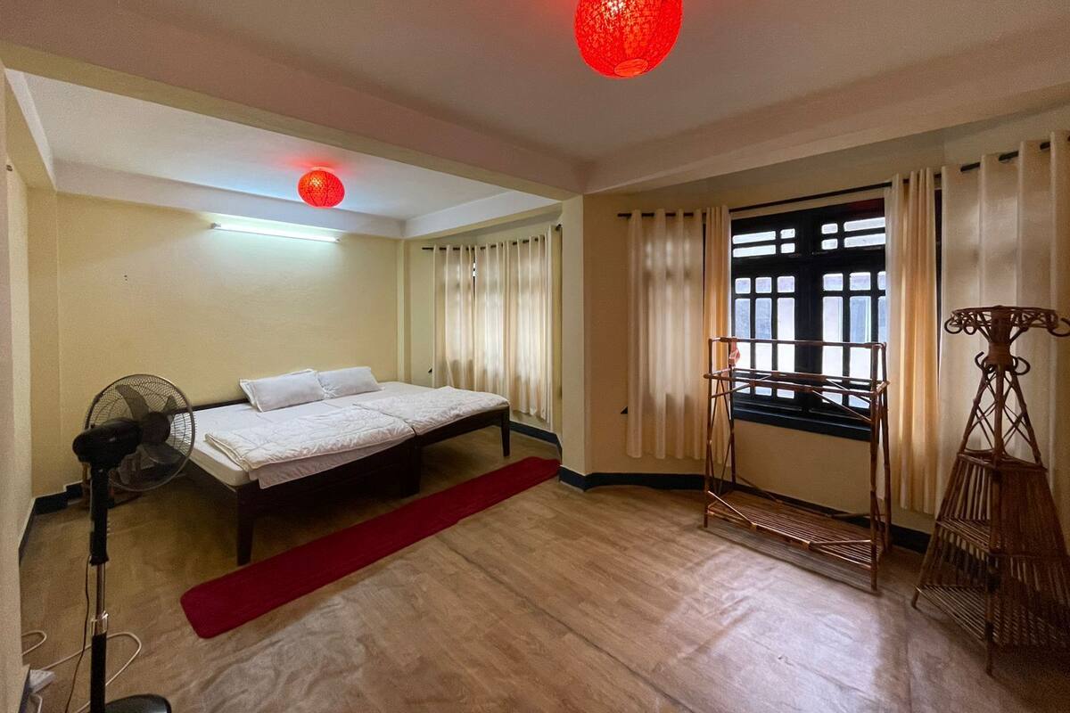 A room with a bed, a standing fan, a red rug, a window with curtains, and a red hanging light fixture.
