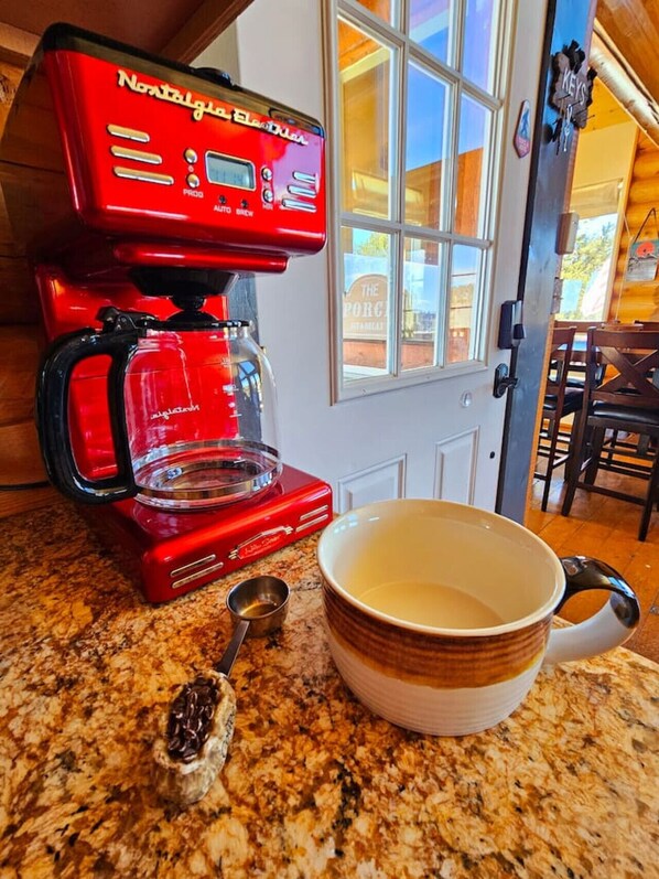 Una cafetera roja sobre una encimera de cocina, con una jarra de vidrio transparente, una cuchara medidora y una taza.