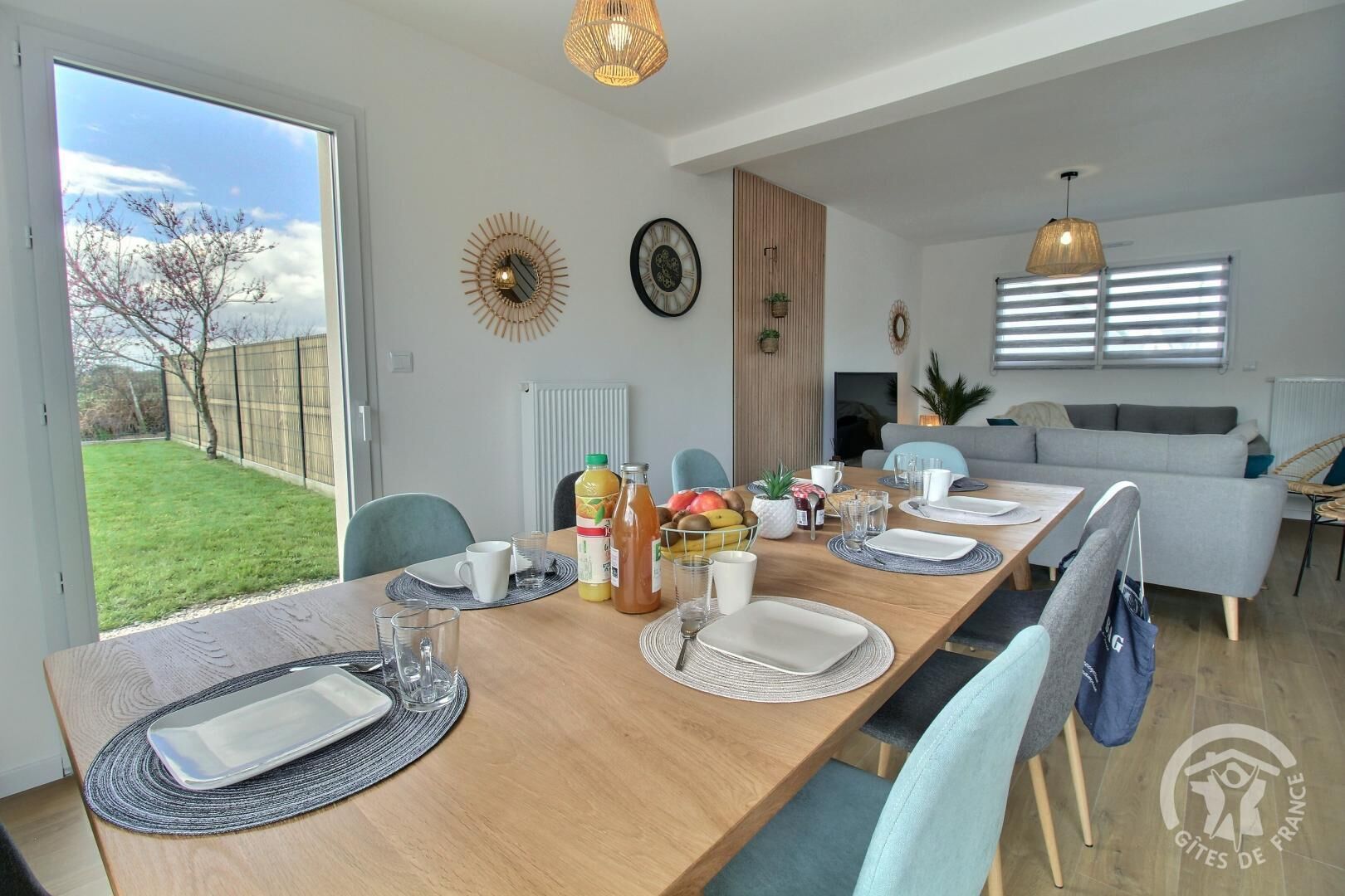 Un espace de salle à manger moderne, avec une table en bois dressée pour quatre personnes, entourée de chaises. On y trouve des horloges décoratives murales, une grande fenêtre avec des stores et une vue sur un jardin verdoyant.