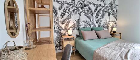 A bedroom with a bed, a wooden desk, a chair, a nightstand, a lamp, and a wall with a tropical leaf mural.