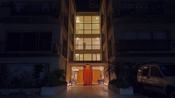 Un edificio de varios pisos con una entrada central, un automóvil estacionado a la derecha y una fachada bien iluminada.