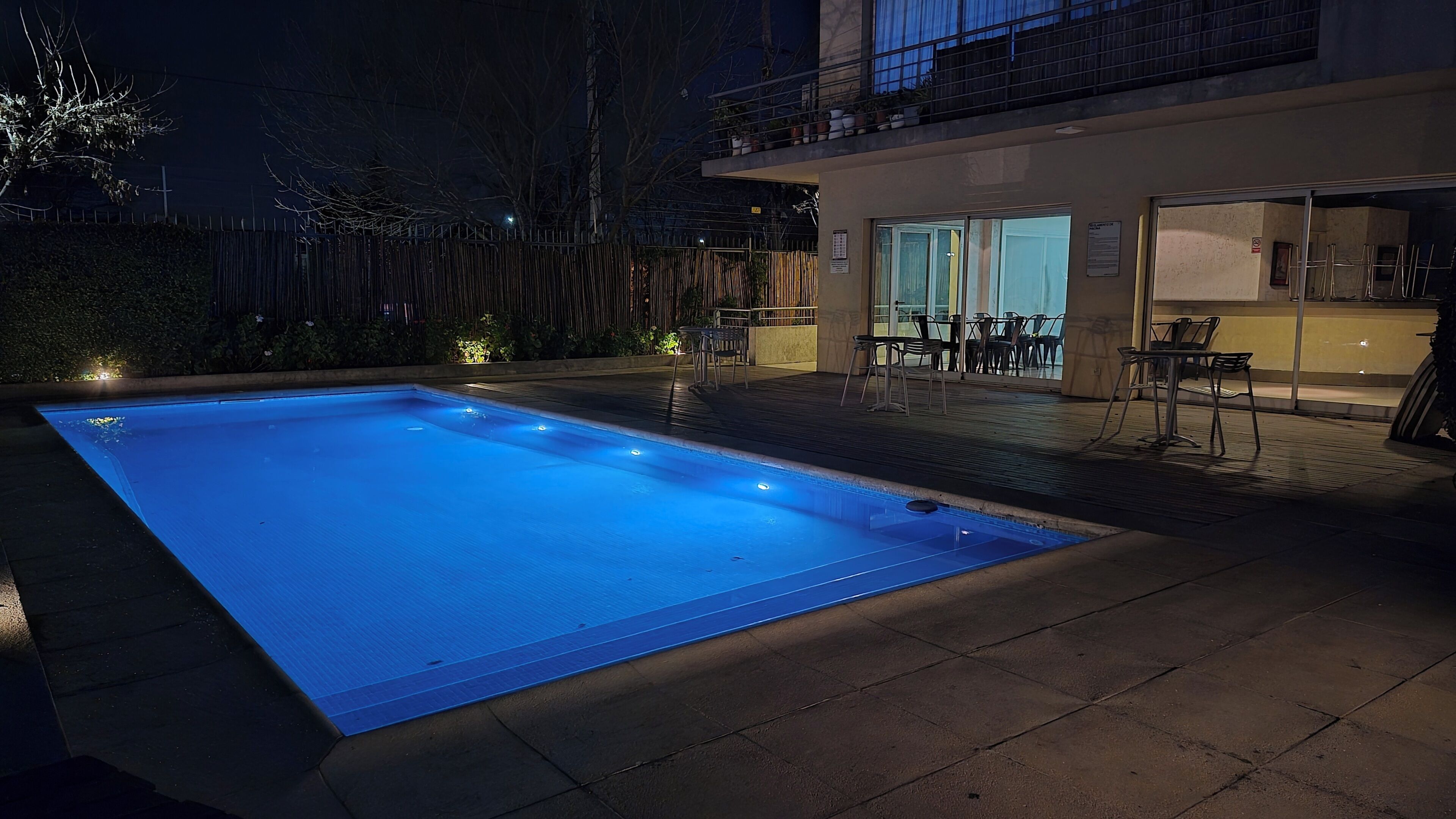 A rectangular swimming pool with a tiled deck surrounding it, a covered outdoor seating area with chairs, and a building with a balcony in the background.