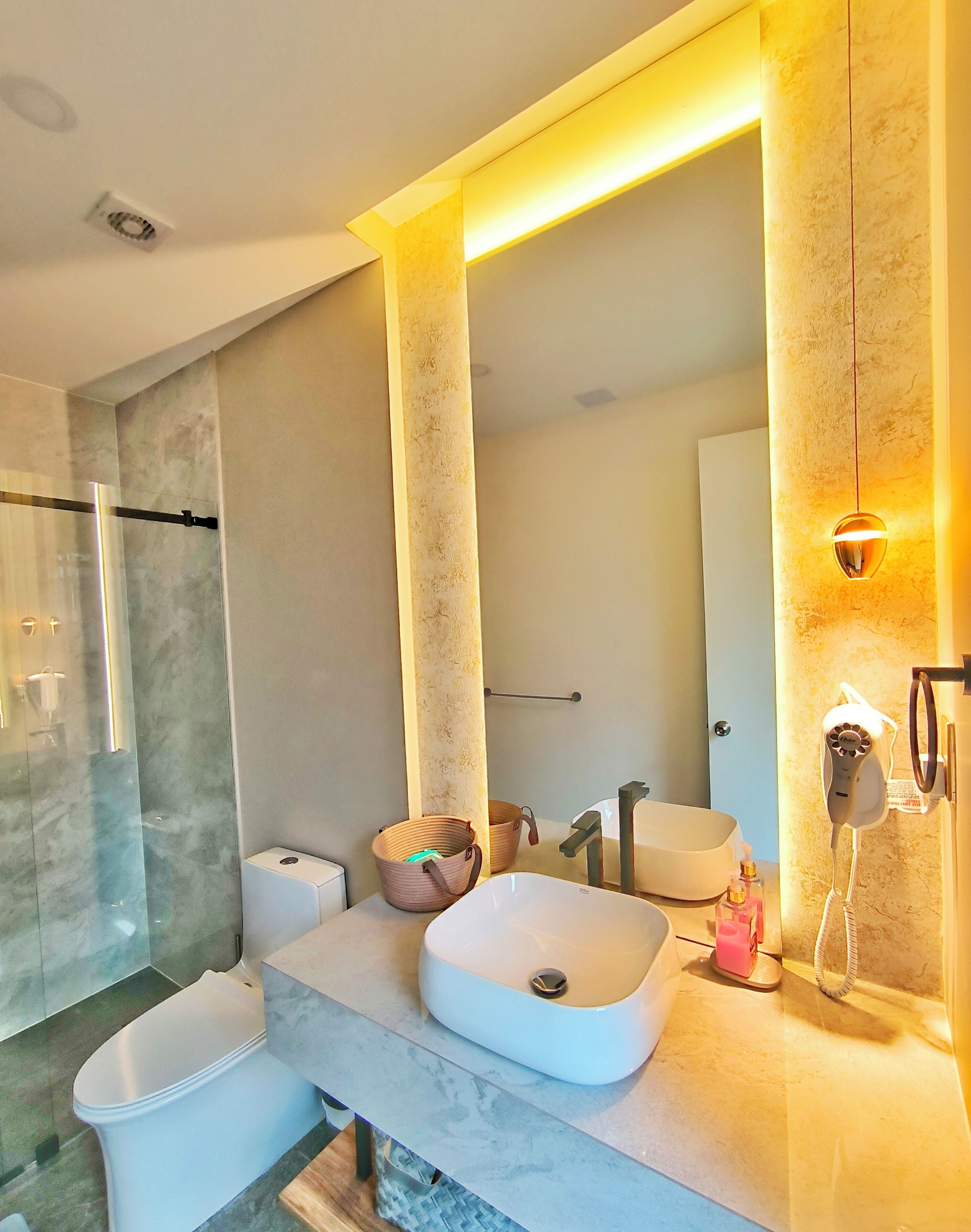 Un baño moderno con un espejo grande, un lavabo blanco, un cabezal de ducha de mano y una ducha con mampara de vidrio.