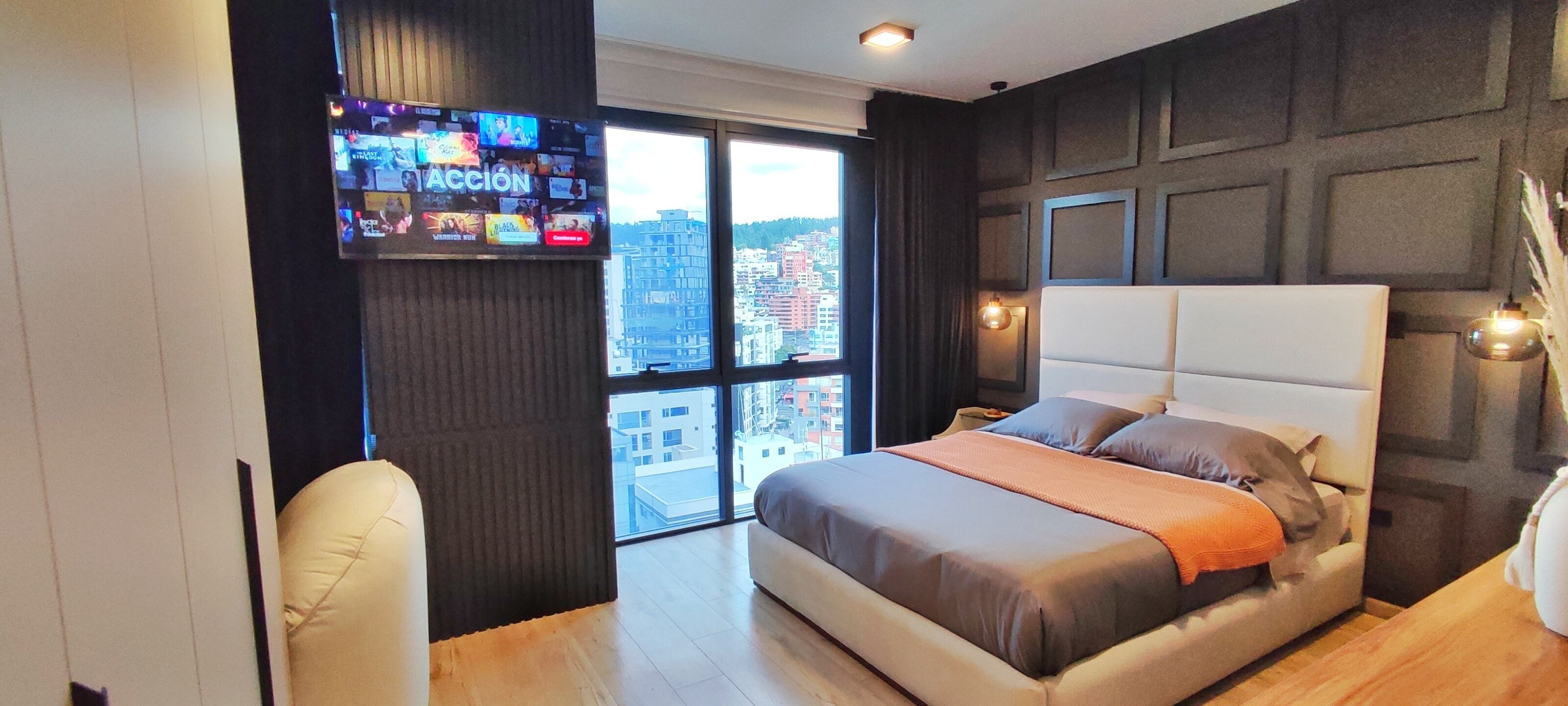 A modern hotel room with a large bed, a TV mounted on the wall, and a view of the city through the window.