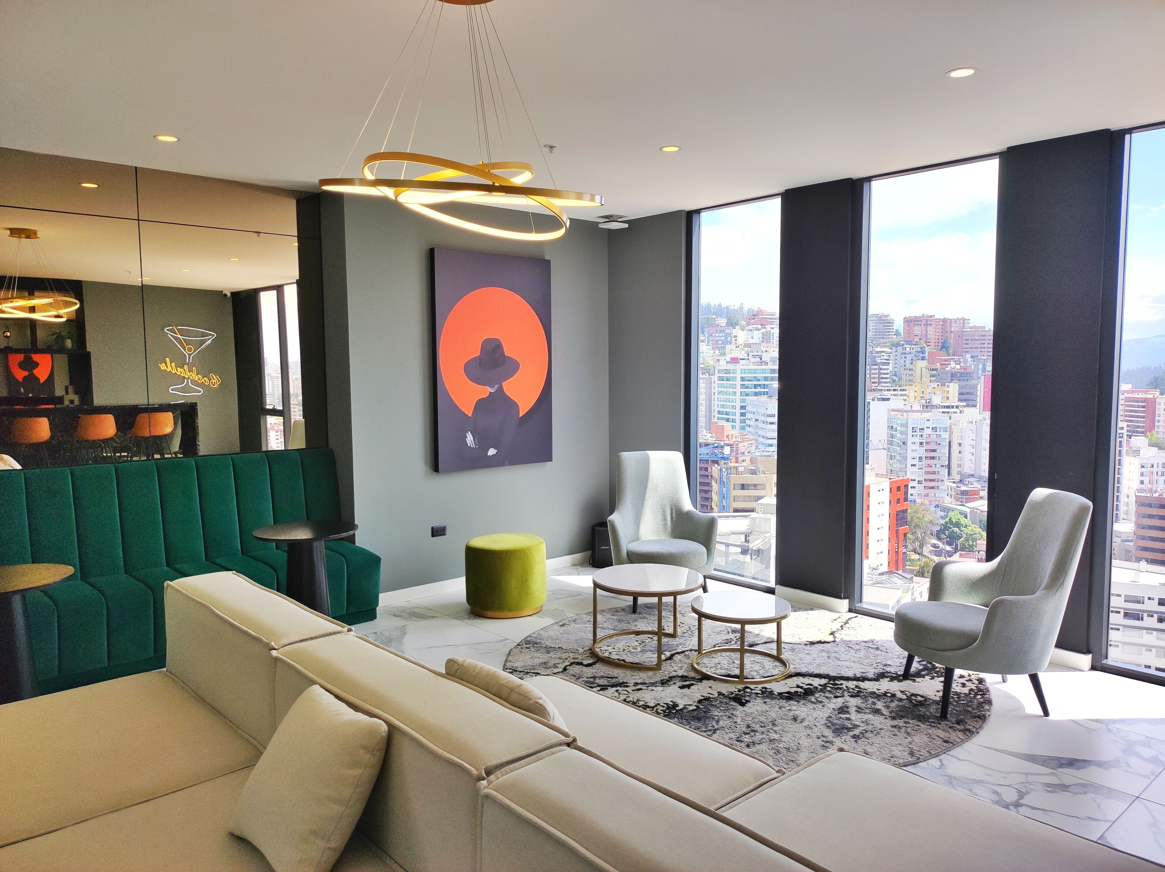 A modern living room with a green sofa, a beige sofa, two armchairs, a round coffee table, and a large window offering a city view.
