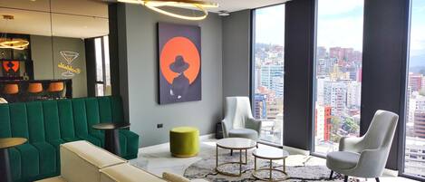A modern living room with a green sofa, a beige sofa, two armchairs, a round coffee table, and a large window offering a city view.
