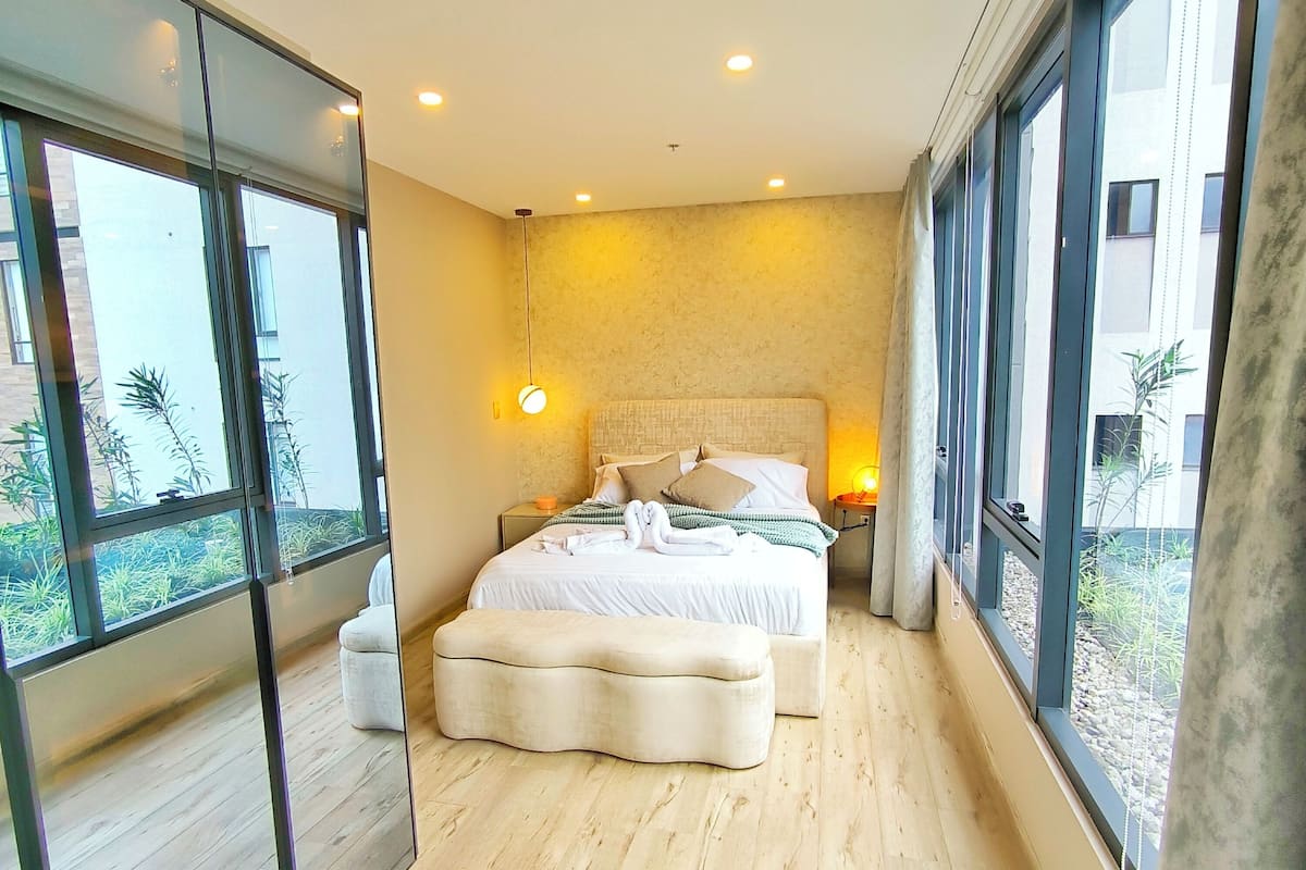 A modern hotel room with a large bed, a sitting area, and large windows offering a view of the outdoors.