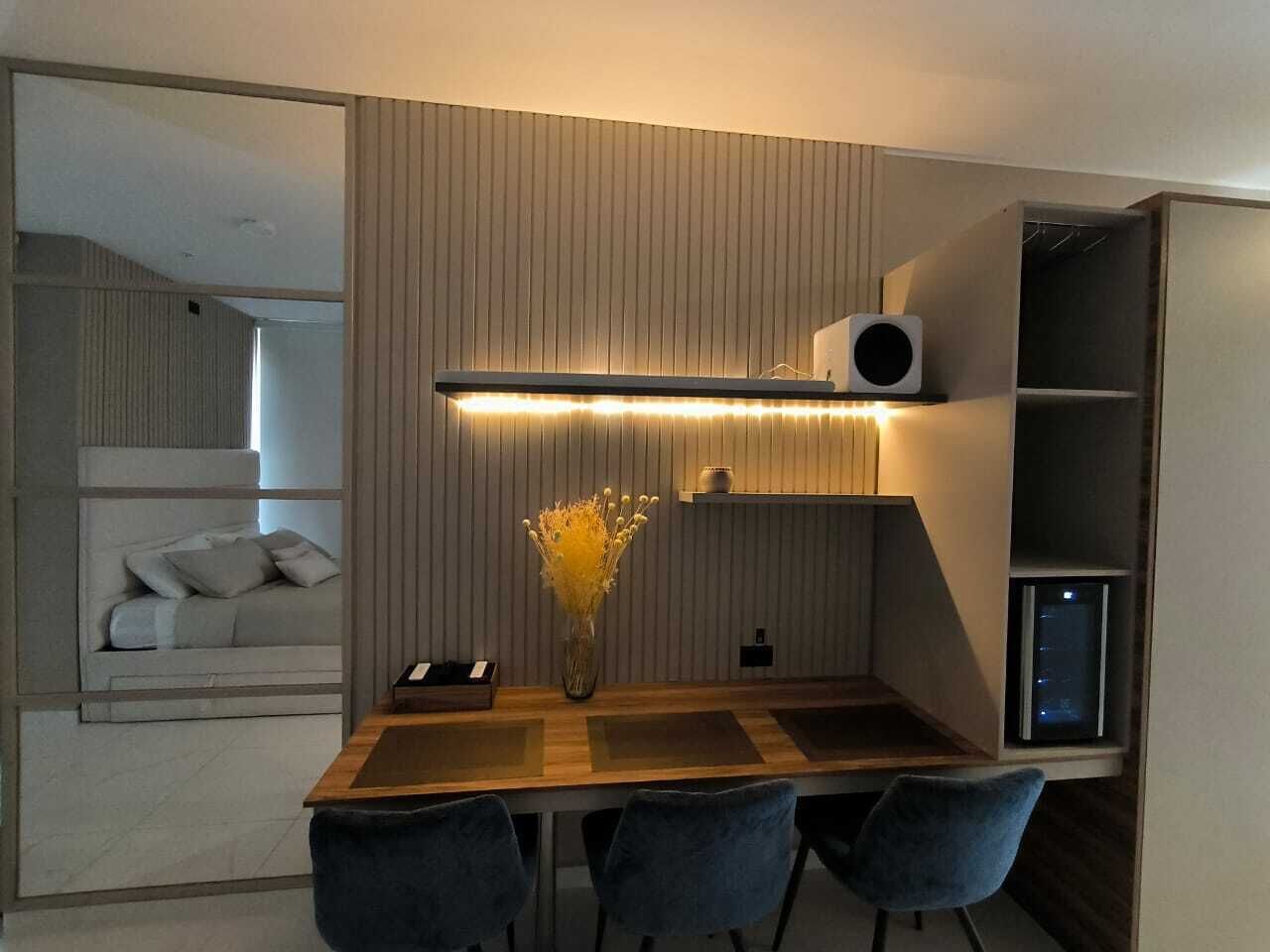 A modern room with a wooden dining table, blue chairs, a vase with yellow flowers, a built-in shelf with a speaker, and a cozy seating area with cushions.