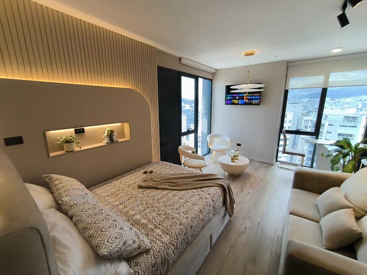 Un dormitorio moderno con una cama grande, una zona de estar con sofá y sillones, un televisor montado en la pared y vistas a la ciudad a través de amplios ventanales.