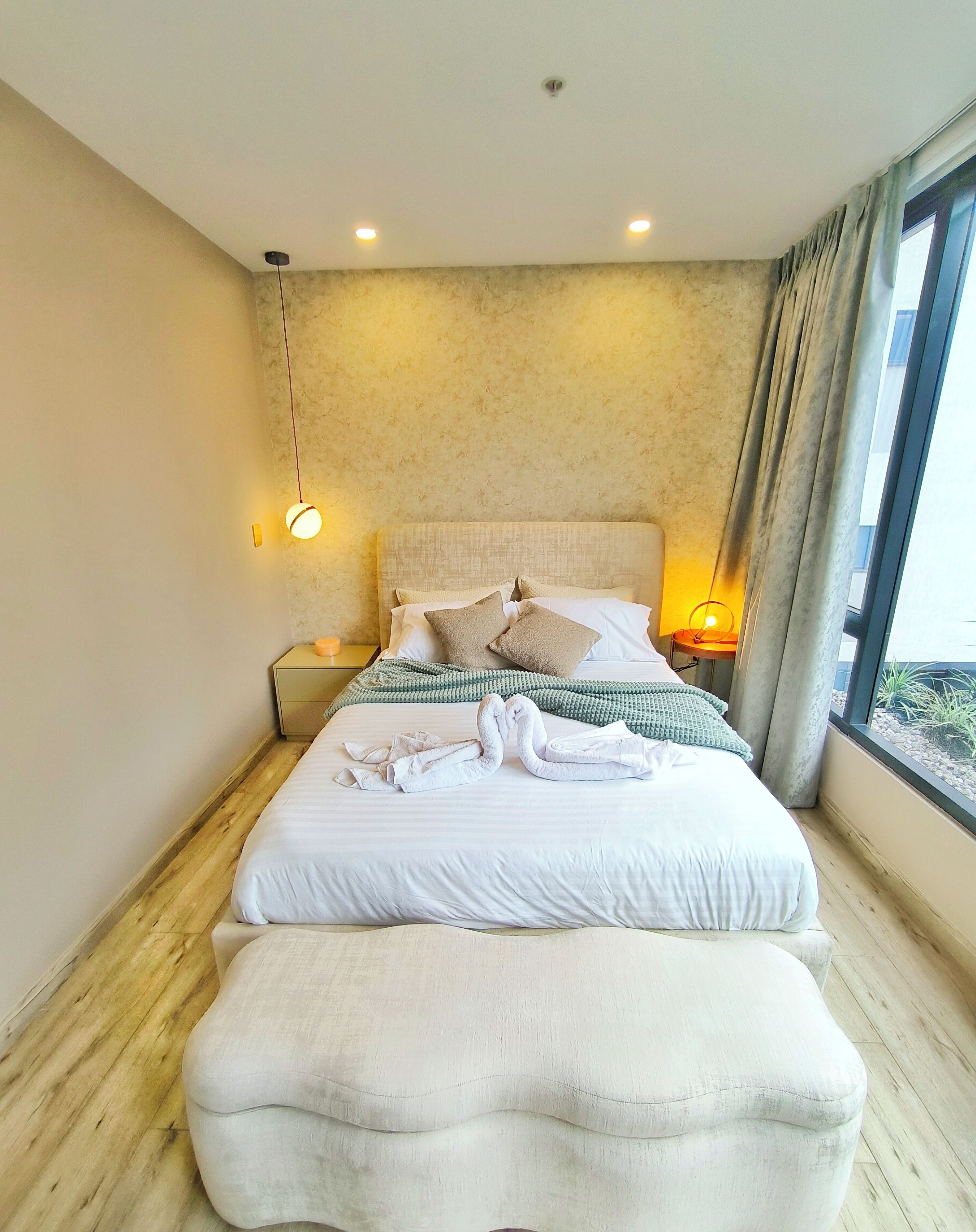 A bedroom with a large bed, a bedside table with a lamp, and a view of the outdoors through a window.