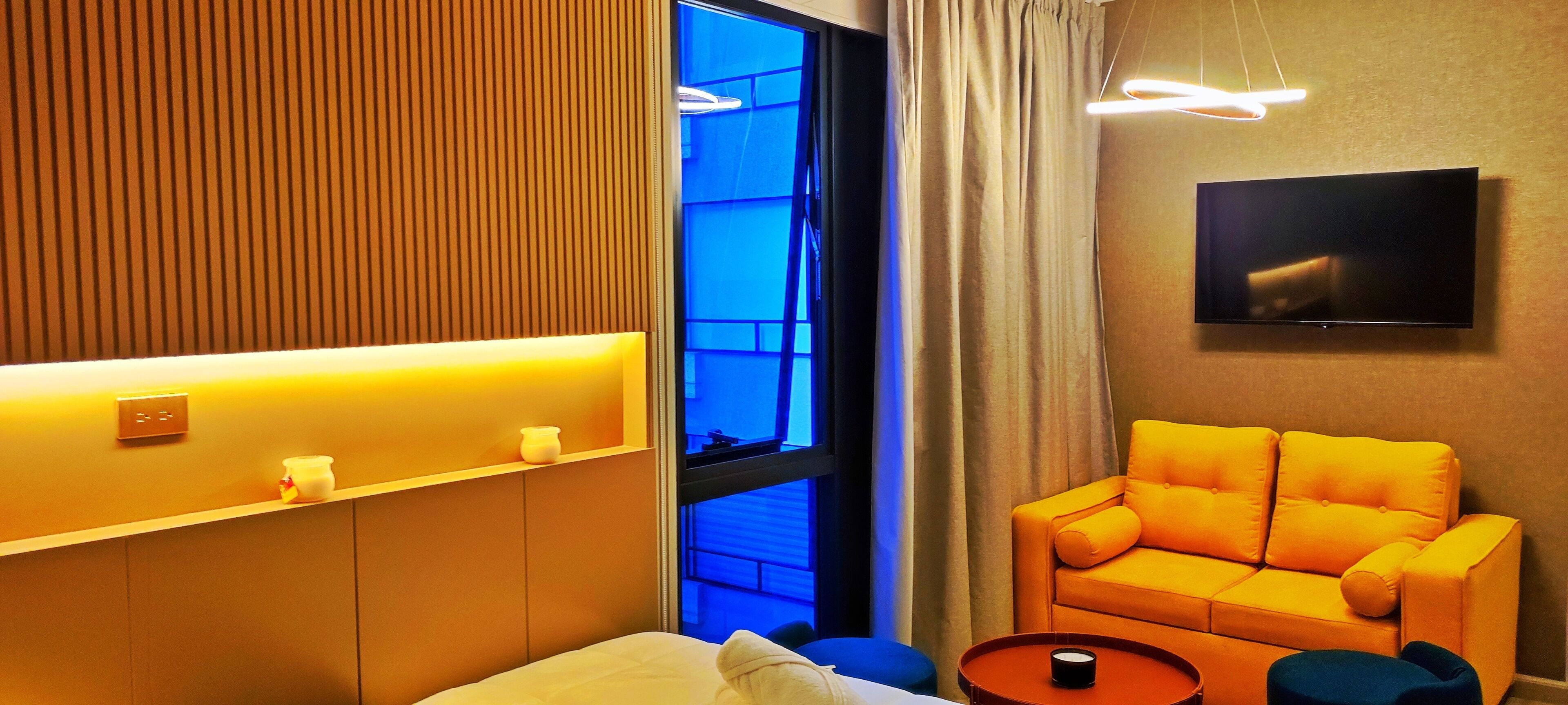 Una habitación de hotel moderna con sofá, una mesita, televisor montado en la pared y ventana con cortinas.