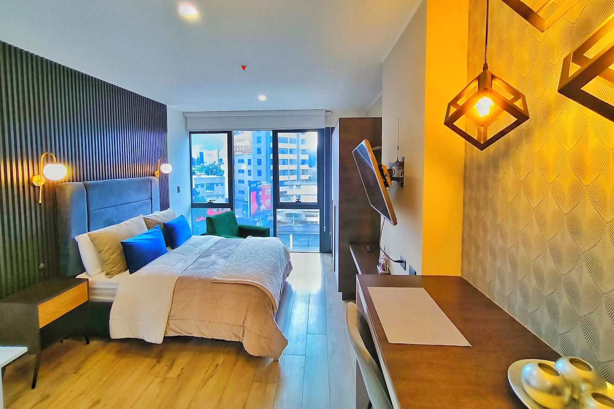 A modern hotel room with a large bed, a desk, a flat-screen TV, and a city view.