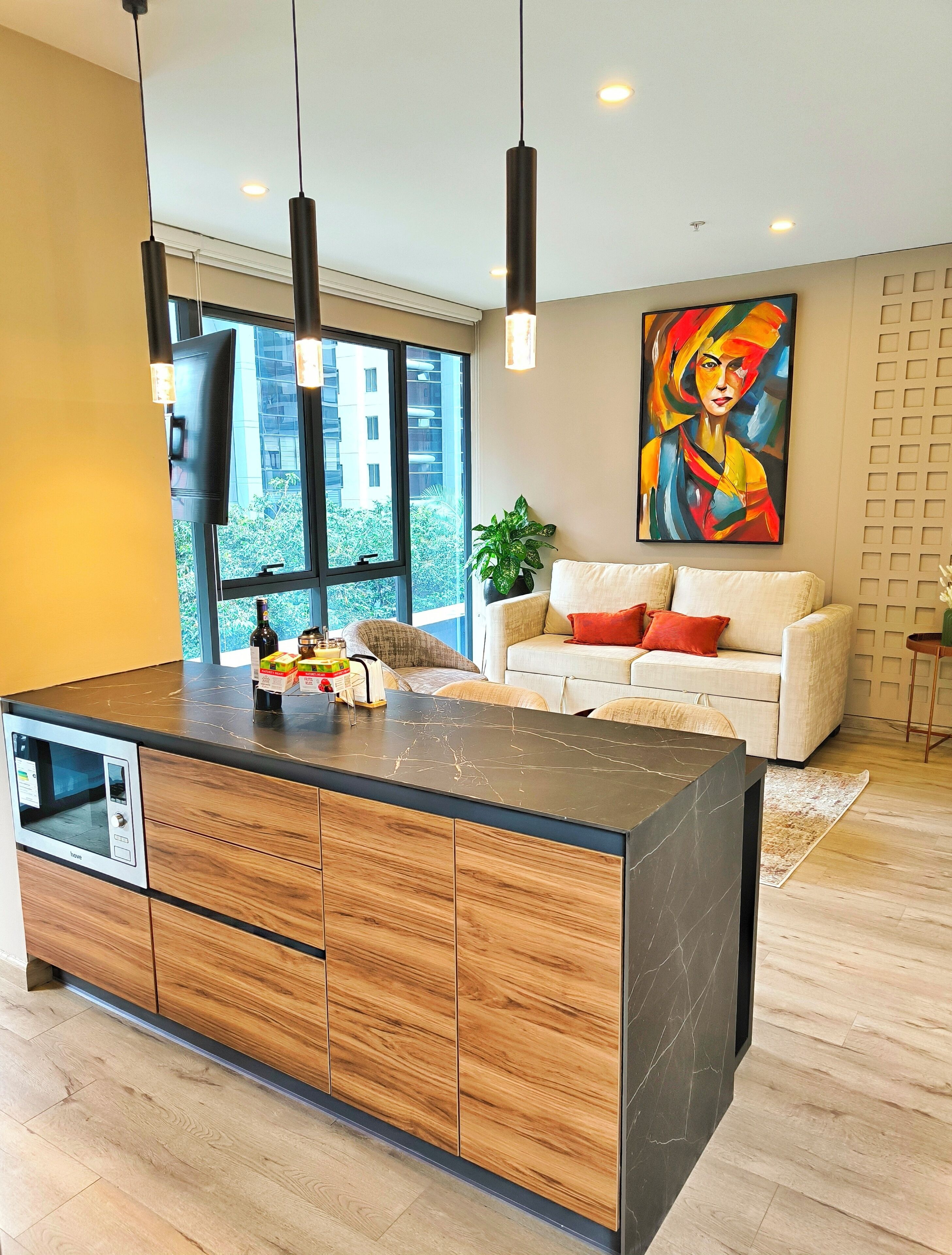 A modern kitchen with a large island, wooden cabinets, and a built-in microwave. There is a sofa and a colorful painting in the living area.