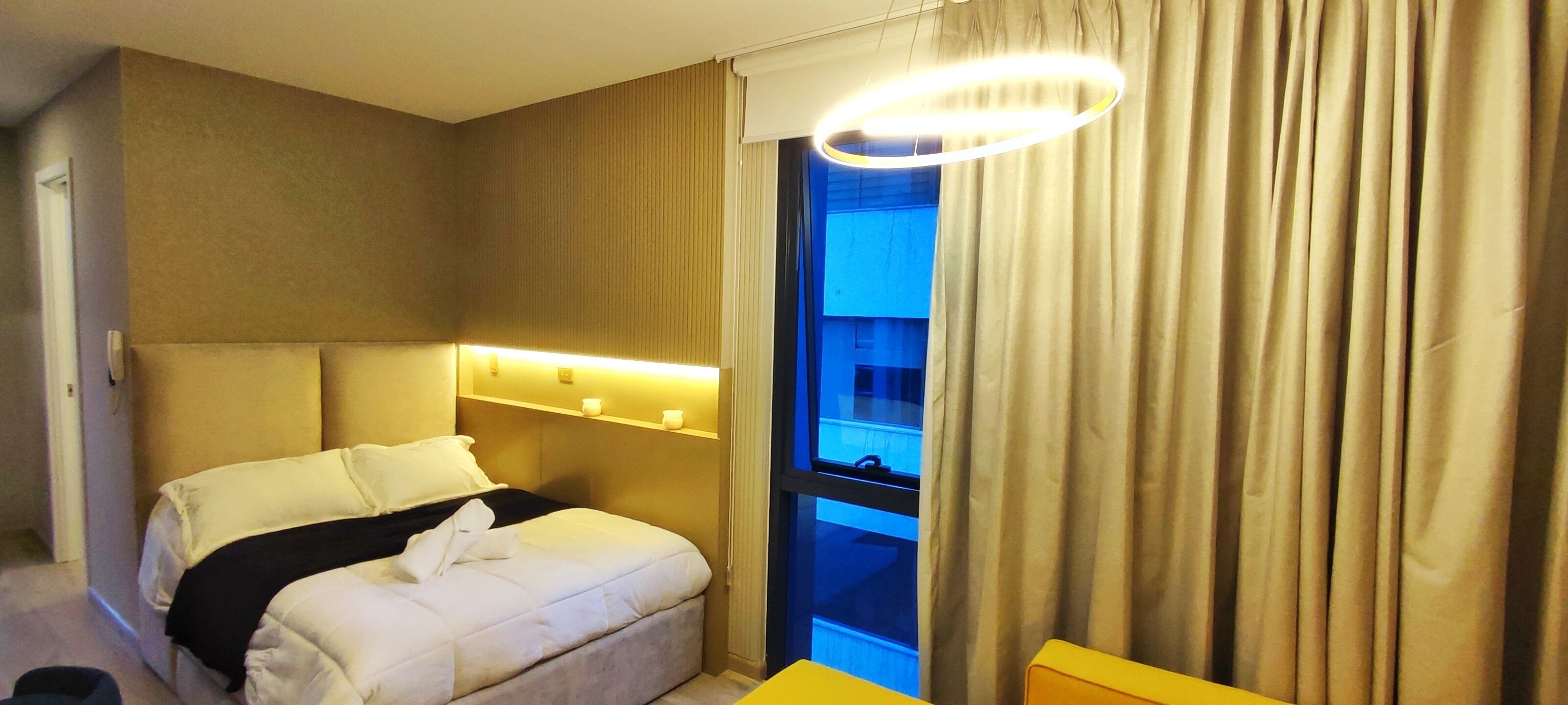 A hotel room with a bed, a bedside table, a window with curtains, and a wall-mounted light fixture.