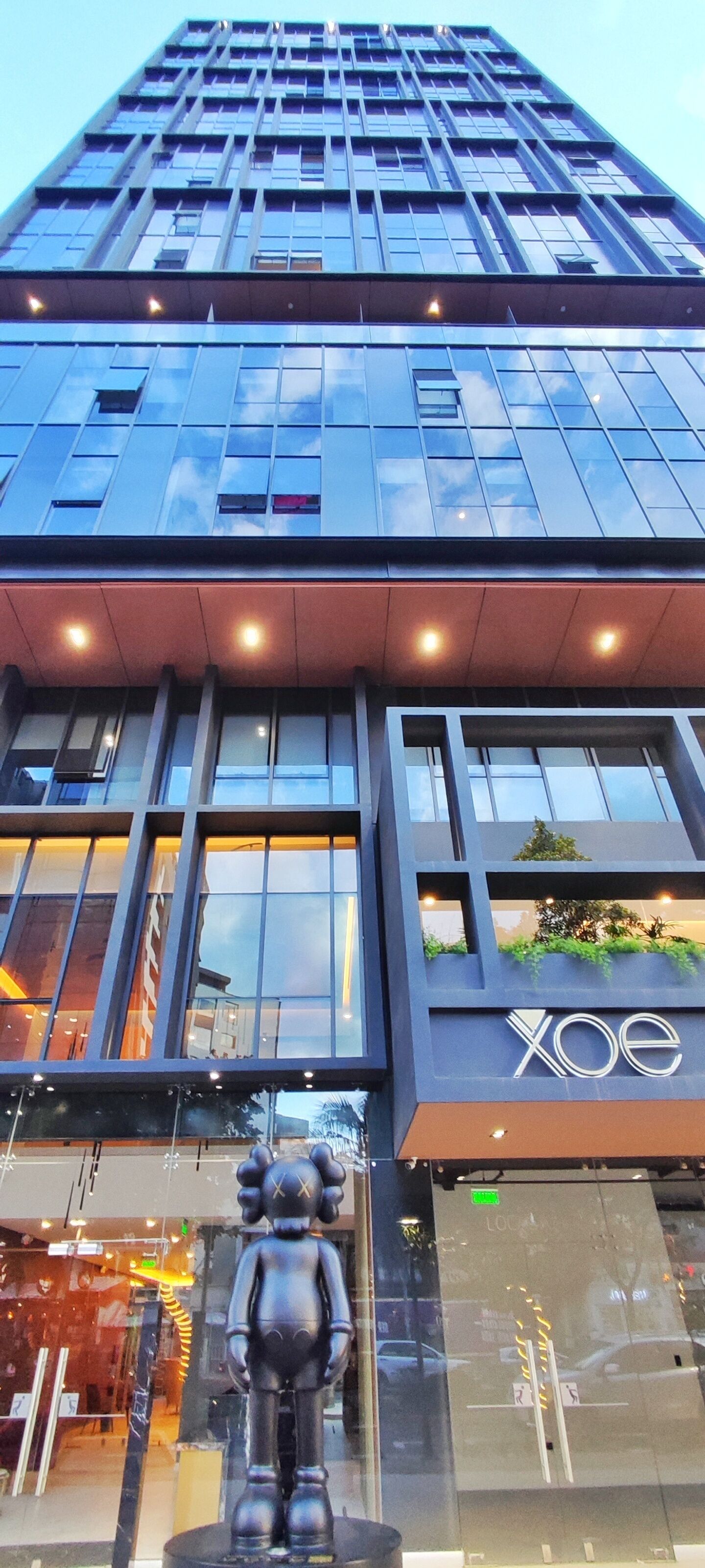 Un edificio moderno con fachada de vidrio, un llamativo letrero 'XOE' y una gran escultura en su parte frontal.