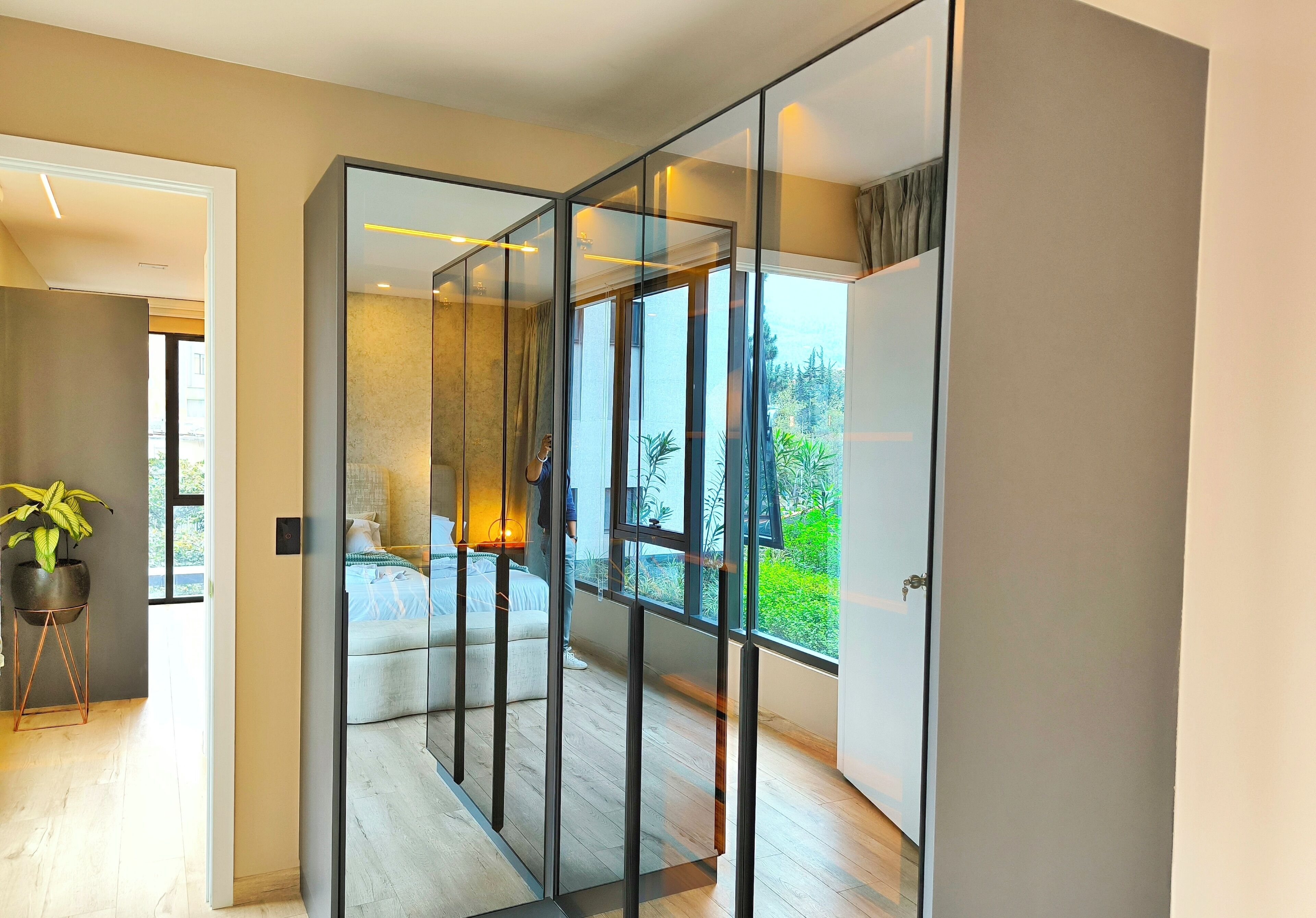 A modern interior with a glass partition, a bed with a blue headboard, and a view of greenery outside.