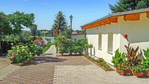 Un chemin pavé mène à un bâtiment blanc surmonté d’un toit rouge, entouré d’une végétation luxuriante et de plantes en pot.