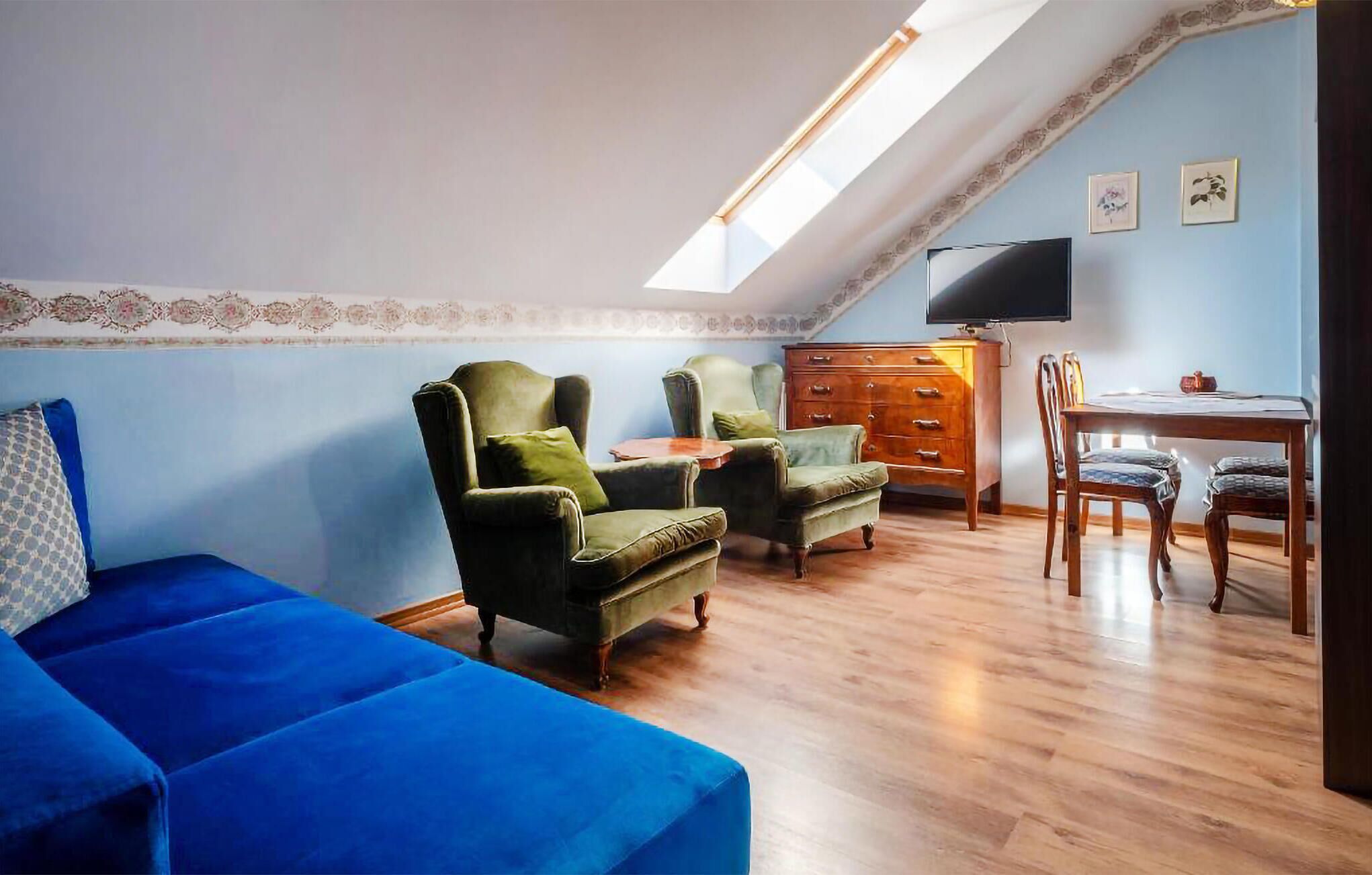 Una sala acogedora con un sofá azul, dos sillones verdes, un aparador de madera, una mesa de comedor con sillas y un televisor montado en la pared.