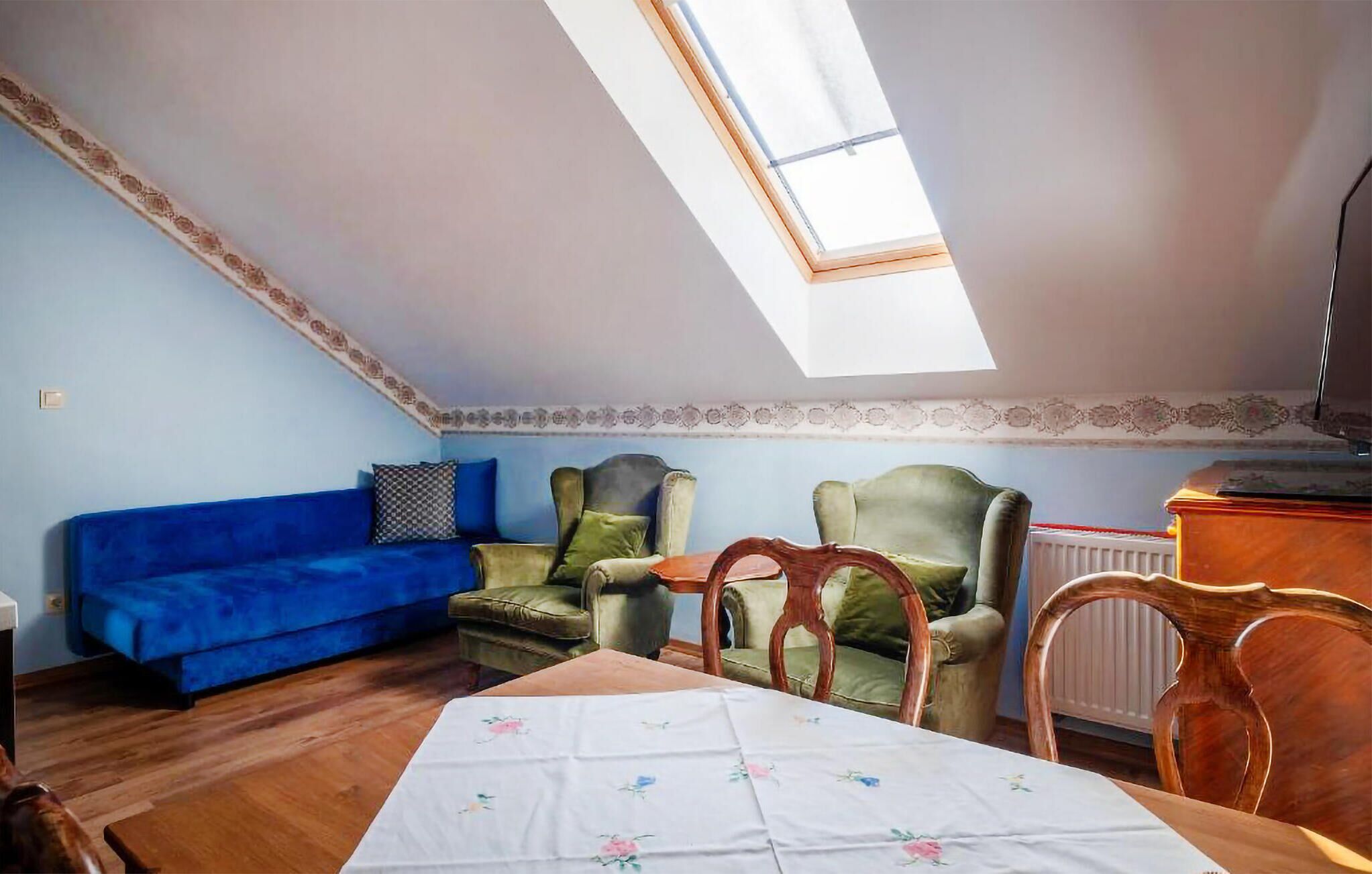 A room with a blue sofa, green armchairs, a wooden table with a floral tablecloth, and a skylight.