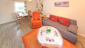Una sala de estar con un sofá, un sillón y una mesa con un florero. Se ve una ventana y una estantería al fondo.