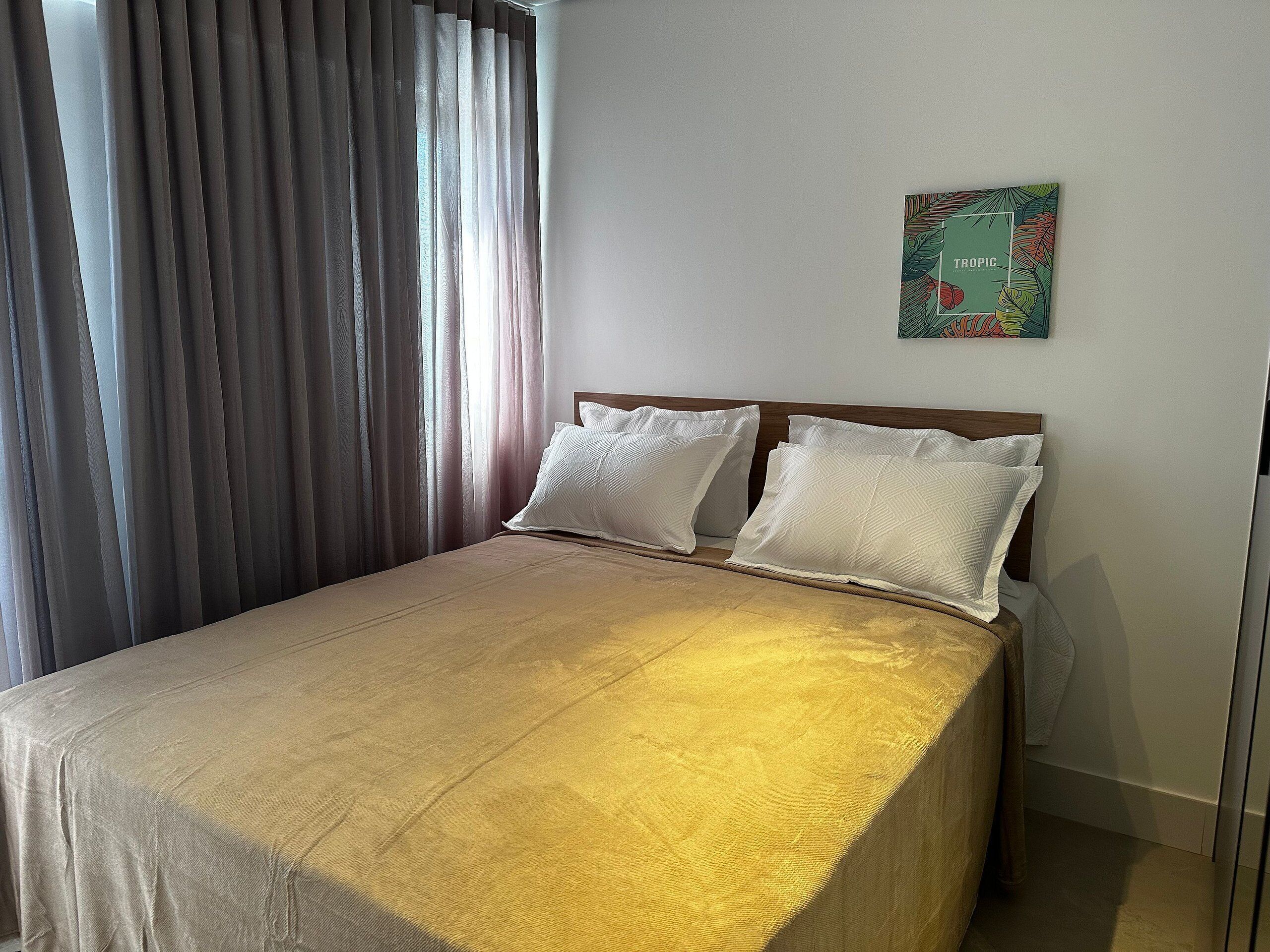A neatly made bed with a beige bedspread and two pillows, a wooden headboard, a window with sheer curtains, and a tropical-themed wall art piece.