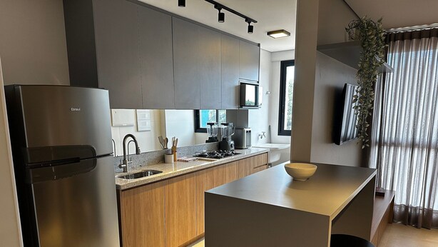 A modern kitchen with a stainless steel refrigerator, wooden cabinets, a built-in microwave, and a large island with stools. - PARK SUL - Edifício Essence (Guará)