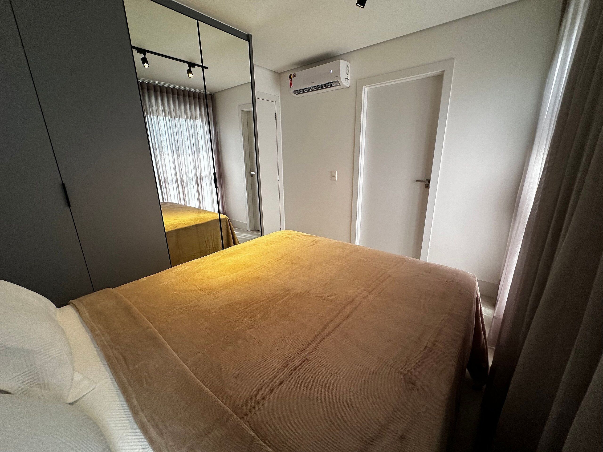 A hotel room with a bed, a wardrobe, a mirror, an air conditioning unit, and a door.