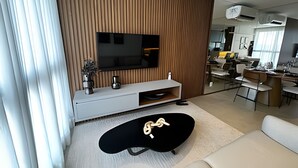 A modern living room with a flat-screen TV, a white entertainment unit, a black coffee table, and a sofa. There is a kitchen area visible in the background. - PARK SUL - Edifício Essence (Guará)