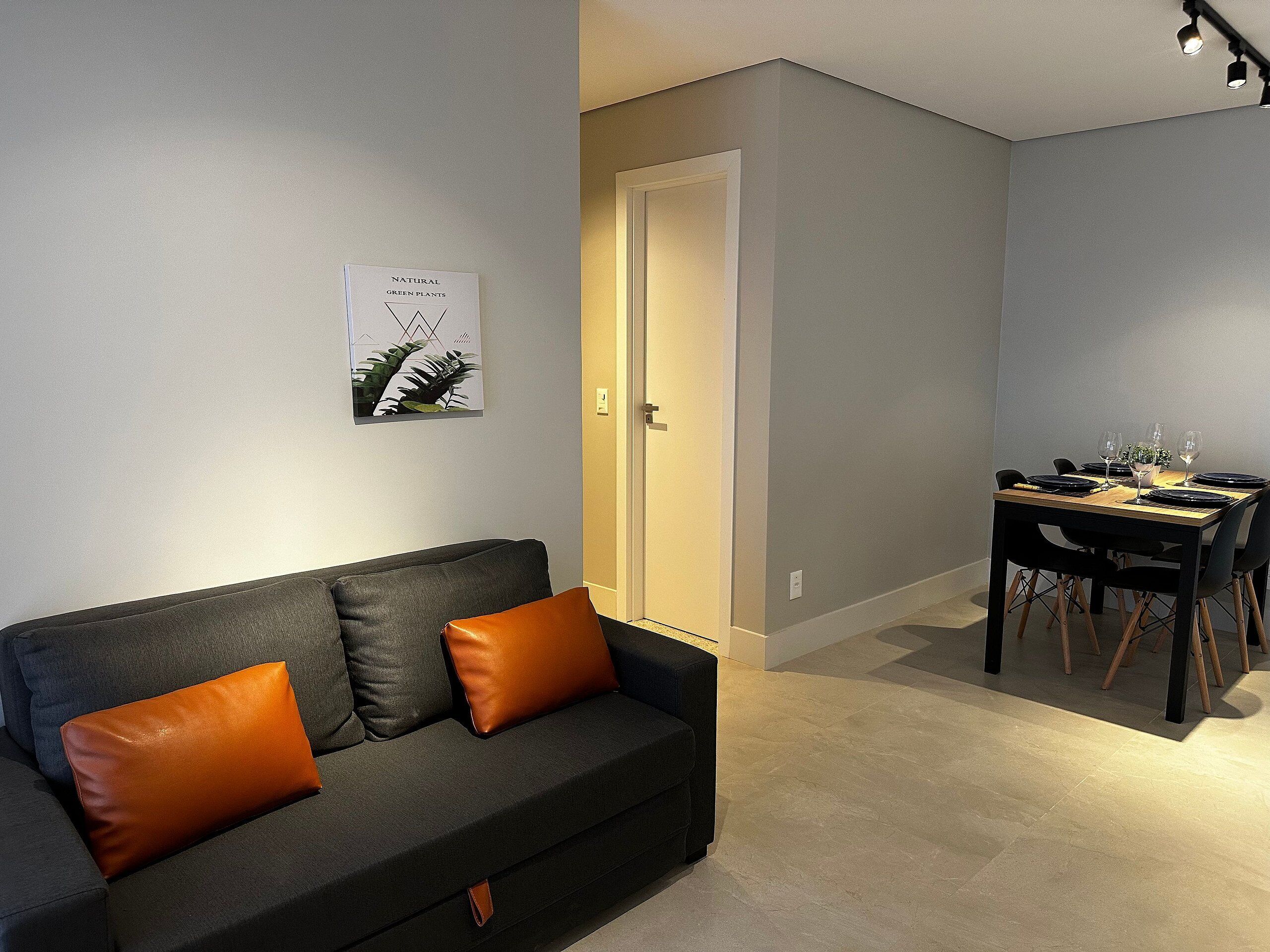 A modern living room with a dark grey sofa, a brown cushion, a dining table set with plates and glasses, and a framed picture on the wall.