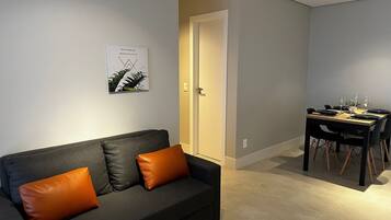 A modern living room with a dark grey sofa, a brown cushion, a dining table set with plates and glasses, and a framed picture on the wall.