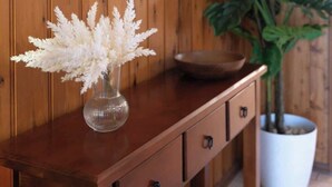 Een houten console tafel met een vaas witte bloemen, een grote potplant en een spiegel met gouden lijst.