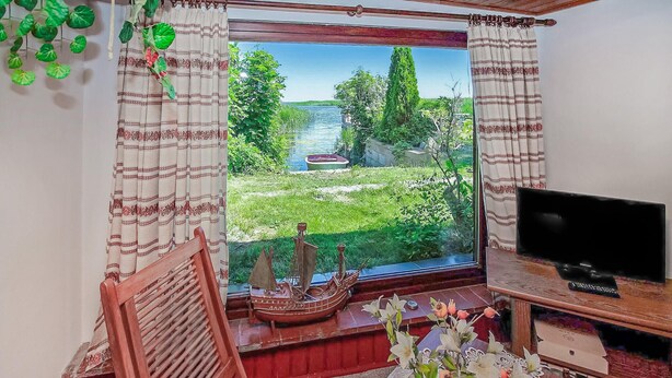 Een kamer met een groot raam met uitzicht op een tuin en een rivier. Er staat een houten stoel, een klein tafeltje met een bloevas en een televisie op een houten plank.