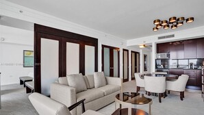 Un salon moderne avec un canapé, des fauteuils et une table basse en verre. On y trouve également un coin cuisine doté de meubles sombres et d’un four à micro-ondes.